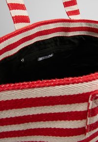 Sac fourre-tout rayé avec des motifs rouges et crème, en matériau tissé, et un intérieur en tissu noir avec une poche zippée et une étiquette.