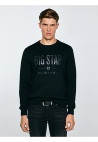 Jauns vīrietis melnā džemperī ar uzrakstu "BIG STAR Your Life. Your Jeans.", kombinācijas melnas džinsu bikses un melns josta, stāv neitrāli.