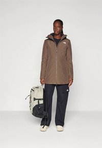 Brązowa wodoodporna kurtka z zamkiem błyskawicznym, kapturem i bocznymi kieszeniami; w połączeniu z czarnymi spodniami i szarym plecakiem z regulowanymi paskami.