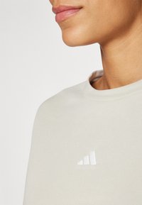 Šviesiai smėlinis džemperis su apvaliu kaklu, kuriame yra baltas Adidas logotipas. Medžiaga atrodo minkšta ir tekstūruota.