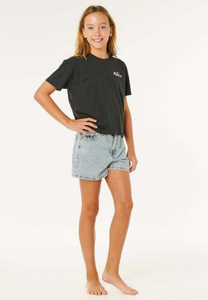 Jong meisje staat op blote voeten, draagt een zwart t-shirt en lichtblauwe denim shorts, glimlacht met één hand op haar heup.