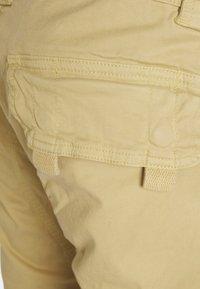 Beige cargo pants with a textured fabric, featuring a large side pocket and reinforced stitching along the seams.