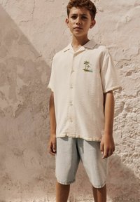 Chemise crème à manches courtes avec broderie de palmier, associée à un short en denim bleu clair. Le tissu a une finition texturée.