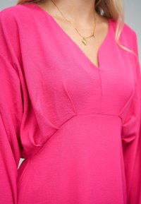 Vaaleanpunainen, väljälahkeinen mekko, jossa on syvä V-pääntie ja pitkät hihat, pehmeä, kevyt kangas ja hienovarainen tekstuuri.