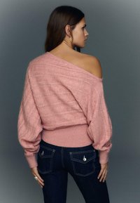 Lyserød strikket bluse med åbne skuldre, tekstureret mønster og balloneærmer, kombineret med mørkeblå jeans med metaldekorerede baglommer.