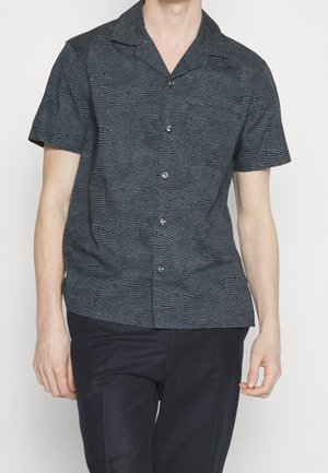 Homme portant une chemise à manches courtes, sombre et à motif, boutonnée, avec un pantalon noir, debout devant un fond uni.