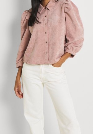 Camisa de pana rosa con botones y mangas abullonadas, combinada con pantalones blancos de cintura alta; presenta botones visibles y una textura suave.