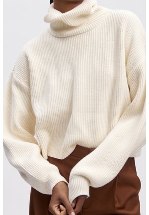 Personne portant un pull à col roulé côtelé crème avec des manches surdimensionnées et un pantalon marron, sur un fond clair uni.