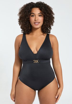 Kobieta z kręconymi włosami w czarnym jednoczęściowym kostiumie kąpielowym z głębokim dekoltem w kształcie litery V i złotym łańcuszkiem w talii, stojąca na jednolitym tle.