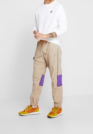 Joggers en nylon beige avec des empiècements violets et un cordon de serrage orange. Associés à des baskets jaunes et rouges. Sweat-shirt blanc avec logo noir.