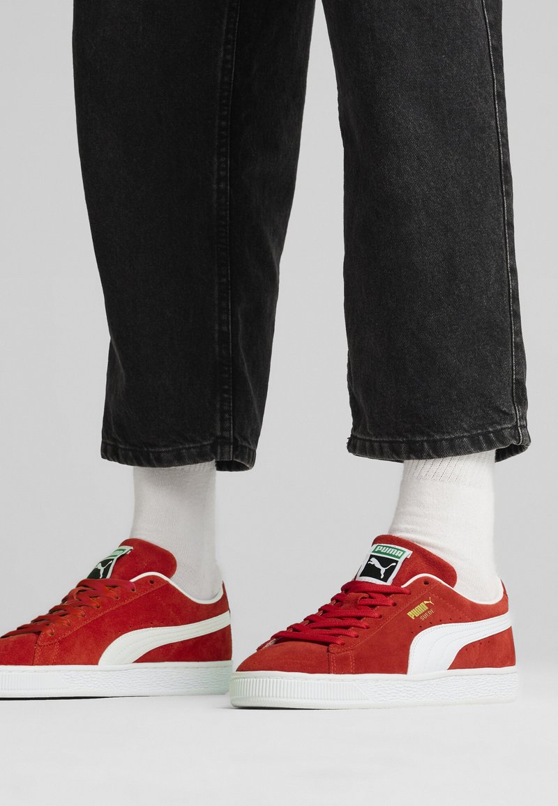 Røde nubuck sneakers med hvide detaljer, gummisål og grøn logo-patch, parret med sorte jeans og hvide sokker.