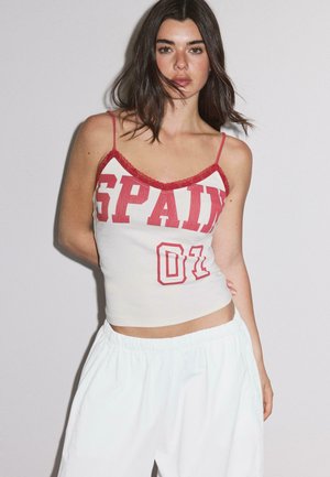 Mladá žena s tmavými vlasmi oblečená v bielom tielku so štíhlymi ramienkami s červeným nápisom „SPAIN 01“ a voľných bielych nohaviciach, stojaca pred jednofarebným pozadím.