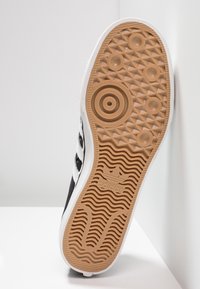 Svart sneaker med vita ränder, texturerad gummisula, cirkel- och diamantmönster samt en gummiyttersula. Har Adidas-logotyp.