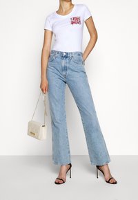 Vit åtsittande t-shirt med grafisk tryck, ljusblå högmidjade jeans, beige vadderad handväska och svarta remsandaler.