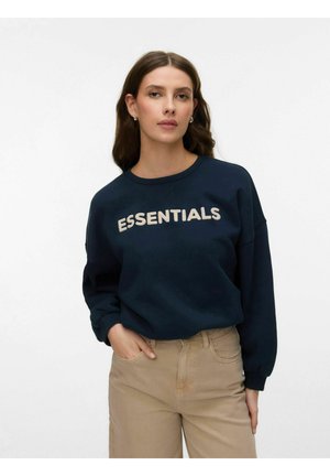 Kvinde med langt brunt hår iført marineblå sweatshirt med teksten "ESSENTIALS" og beige bukser, poserer mod en ensfarvet hvid baggrund.