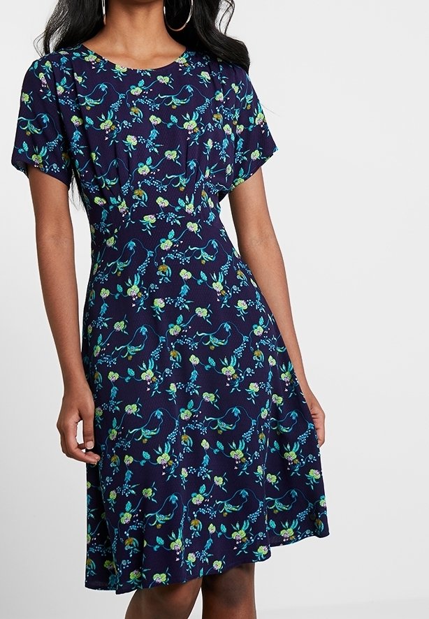 Mekko tummansinisestä kankaasta, jossa on lyhyet hihat ja vihreä- sekä akvamerenvärinen kukkakuosi. Kellotettu muotoilu ja pehmeä kangas, joka valuu kauniisti.