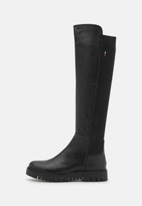 Botas negras de cuero lisas hasta la rodilla, con un panel lateral elástico, suela de goma y detalles de banda texturizada. Presenta un sutil acento de logo.