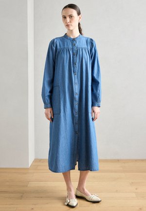 Lys blå denimkjole med høy hals, lange ermer, knappestolpe foran, en samlet kropp og en lomme foran. Brukt med lyse leiligheter.