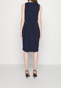 Marinblå ärmlös klänning med en fitted bodice, plisserade detaljer vid midjan och knälång fåll. Bärs med svarta högklackade sandaler.