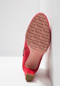 Chaussure à talon haut en daim rouge avec un bout arrondi, fermeture éclair latérale et semelle en caoutchouc marron texturée, ornée du nom de la marque en relief.