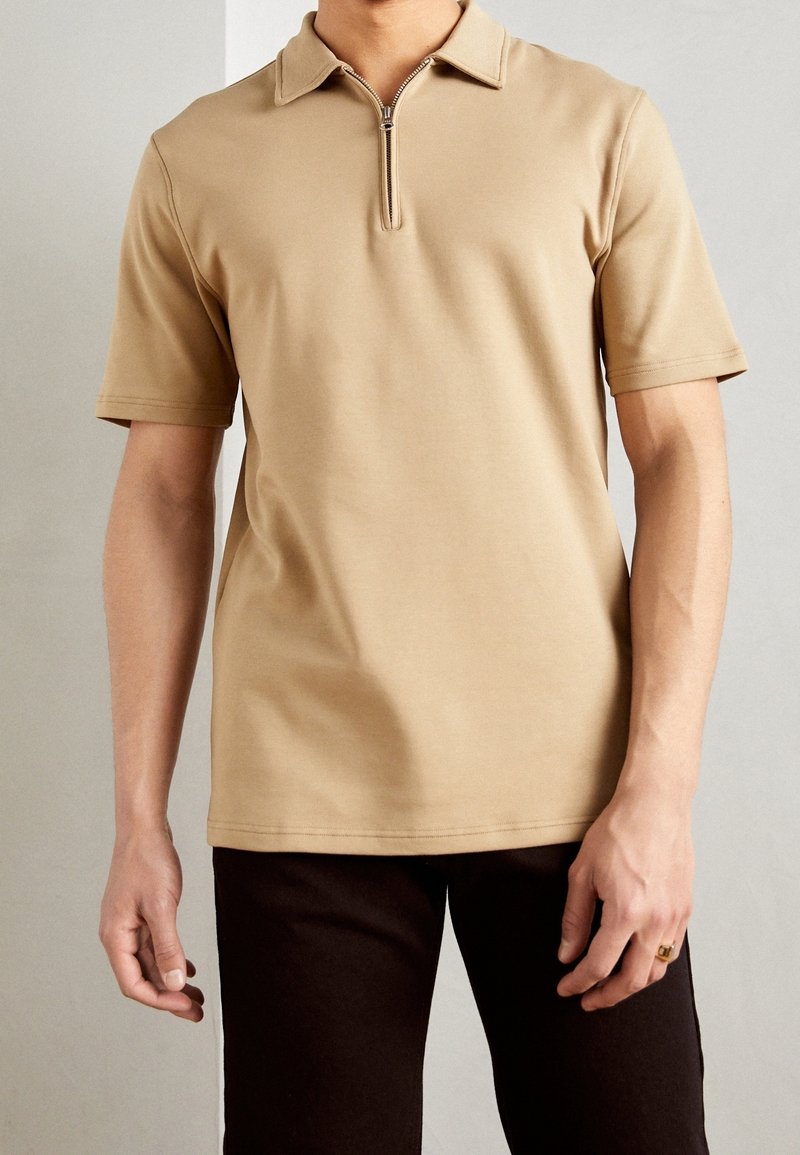 Polo shirt beige à manches courtes avec un col plat et un col zippé, en tissu lisse, associé à un pantalon sombre.