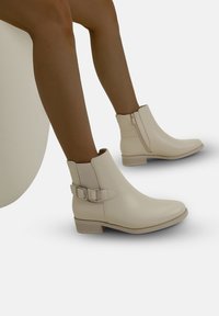 Botas de tobillo beige con un acabado de cuero suave, cremallera lateral y acento de hebilla ajustable, con un tacón bajo apilado.