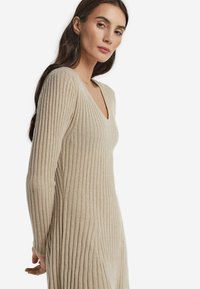 Beiges geripptes Strickkleid mit tiefem V-Ausschnitt und langen Ärmeln. Verfügt über eine figurbetonte Silhouette und weiche Textur, die vertikale Linien betont.