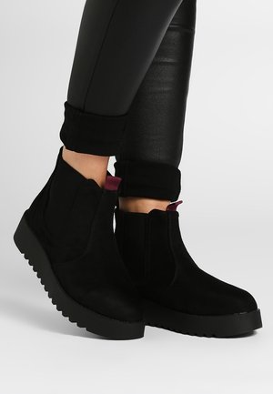 Bottes chevilles en daim noir avec semelles épaisses à reliefs et languettes de tirage rouge foncé, portées avec un pantalon en cuir noir retroussé.