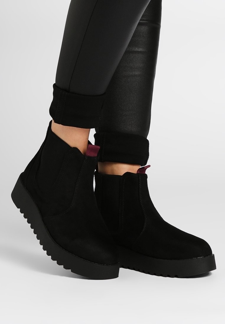 Bottes chevilles en daim noir avec semelles épaisses à reliefs et languettes de tirage rouge foncé, portées avec un pantalon en cuir noir retroussé.