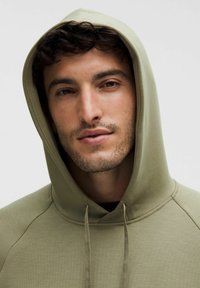Sweat-shirt à capuche vert clair avec une texture lisse, présentant une poche kangourou et un capuchon à cordon. Portrait en gros plan d'un homme le portant.