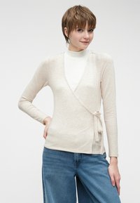 Valkoinen neulepaita, jossa on pehmeä kangas, pitkähihaiset hihat, V-pääntie ja vyötäröllä solmittava yksityiskohta. Käytetty valkoisen poolopaidan päällä.