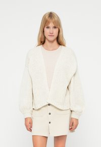 Femme blonde aux cheveux raides portant un gilet en maille crème surdimensionné, un haut beige et une jupe crème boutonnée, debout sur un fond blanc.