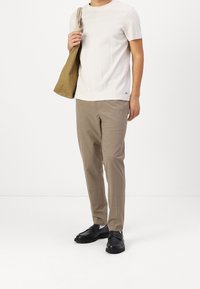 Camicia bianca a maniche corte con texture, pantaloni eleganti beige e scarpe nere. In mano sinistra tiene una borsa a tracolla beige.