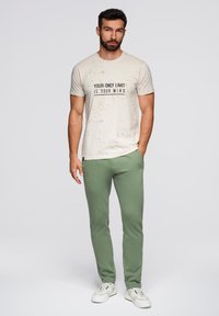 Lichtbeige T-shirt met een grafisch tekstontwerp, gecombineerd met olijfgroene joggers, beide gemaakt van zachte stof. Witte sneakers maken de outfit compleet.