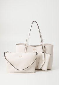 Tři bílé texturované tašky GUESS: velká kabelka s uchy, střední zipová peněženka a malá taštička připojená k kabelce.