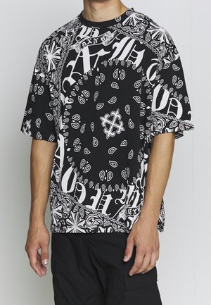 Mann trägt ein schwarz-weißes, bandana-gemustertes oversized T-Shirt und schwarze Cargohosen, steht vor einem einfarbigen Hintergrund.