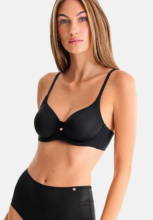 Femme portant un soutien-gorge à armatures noir et une culotte taille haute assortie, debout devant un fond clair uni.
