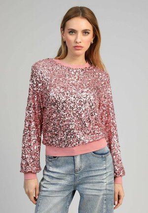 Femme portant un haut à manches longues rose pailleté et un jean bleu clair délavé, debout devant un fond gris uni.