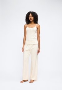 Γυναίκα με κατσαρά μαλλιά που φοράει μπεζ πλεκτό καμησόλ και παντελόνι με φαρδιά πόδια σε απόχρωση που ταιριάζει, όρθια ξυπόλητη μπροστά σε λευκό φόντο.