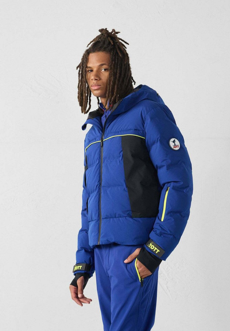 Junger Mann mit Dreadlocks, der eine blau-schwarze isolierte Jacke und passende blaue Hose mit gelben Reißverschluss-Akzenten vor einem schlichten Hintergrund trägt.