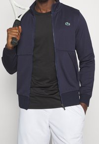 Mörkblå zip-jacka med texturerad yta, med en liten grön krokodil-logotyp på bröstet. Svart t-shirt under, håller en tennisracket.