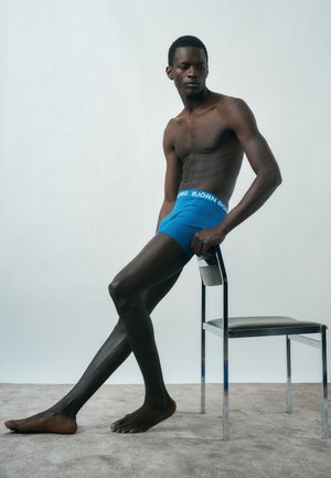 Άνδρας μοντέλο με μπλε μποξέρ Björn Borg, καθισμένος πλάγια σε μεταλλική καρέκλα με μπεζ κάθισμα, μπροστά σε απλό ανοιχτό φόντο.