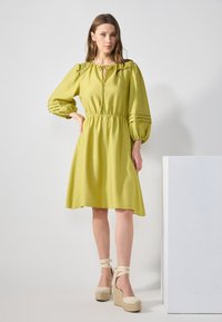 Limegrünes Kleid mit Puffärmel, einem Kordelzug-Ausschnitt und strukturiertem Muster. Kombiniert mit beigen, gestrickten Plateau-Esparille.