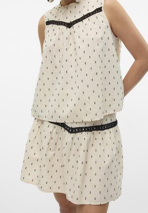 Ensemble top sans manches crème et jupe avec petit motif à pois noirs et bordure en dentelle noire sur la poitrine et la taille, porté par une personne.
