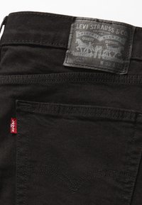 Melnas džinsa bikses ar ādas izšuvumu ar zīmolu, smalkām šuvju detaļām un sarkanu cilpiņu uz aizmugurējā kabatas.