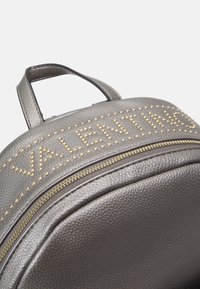 Grijze leren rugzak met goudkleurige studs en "VALENTINO" lettertype, een textuurafwerking en een gouden ritsluiting op het voorvak.