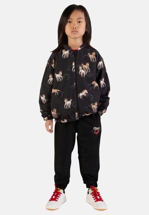 Veste noire à motifs de chevaux colorés, associée à un pantalon de jogging noir avec un logo rouge. Les chaussures sont blanches avec des lacets rouges.