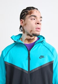 NIKK-jakke i turkis og svart, glidelås lukking, hette-design, stoff med en glatt tekstur, Nike-logo på brystet, lilla skjorte under.