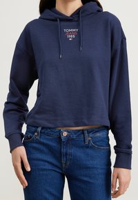 Hoodie cropped blu navy con cordini, con la scritta "TOMMY JEANS 1985" stampata sul petto. Abbinato a jeans di denim blu.