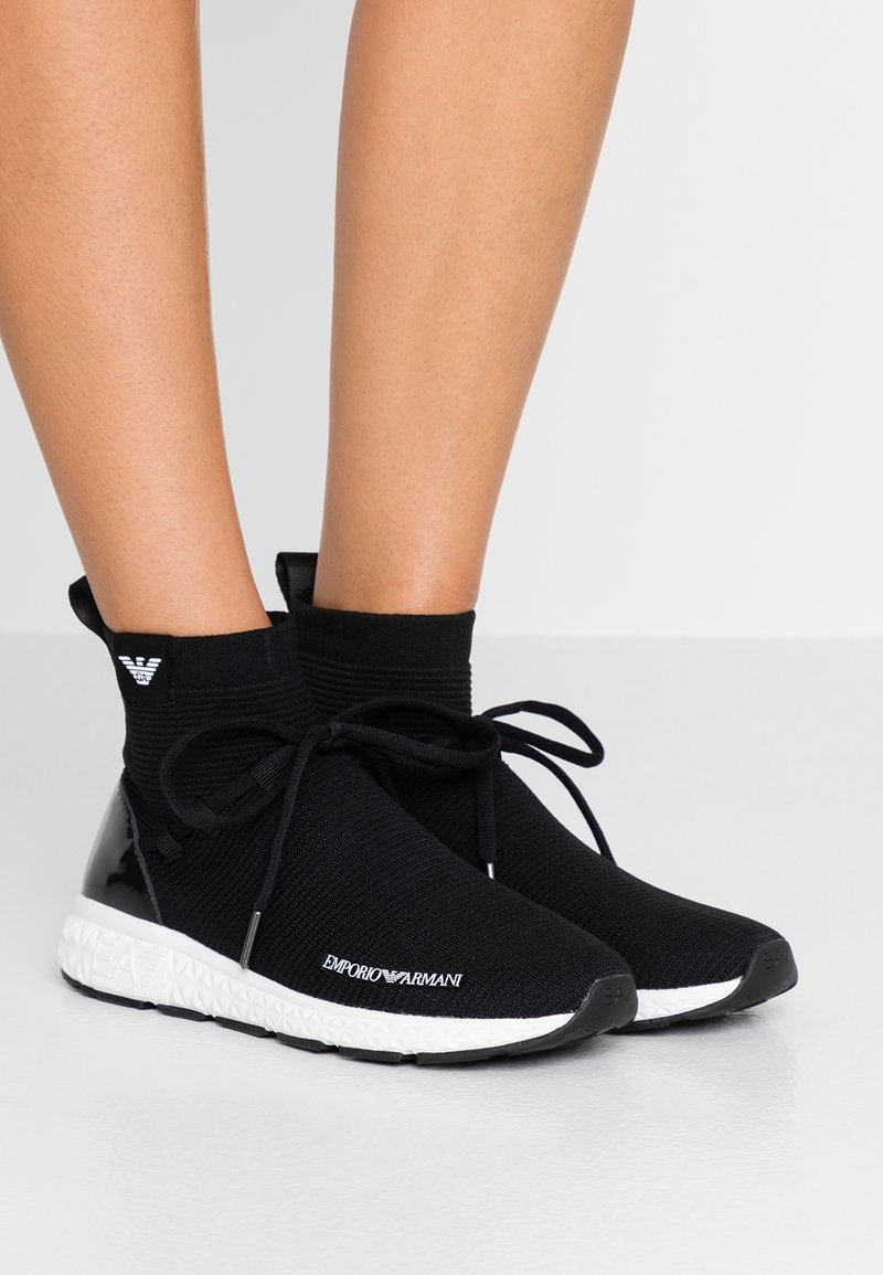 Sneaker alte nere lavorate a maglia con un design elegante e testurizzato. Dotate di un tacco nero lucido e una suola bianca con il marchio "Emporio Armani".